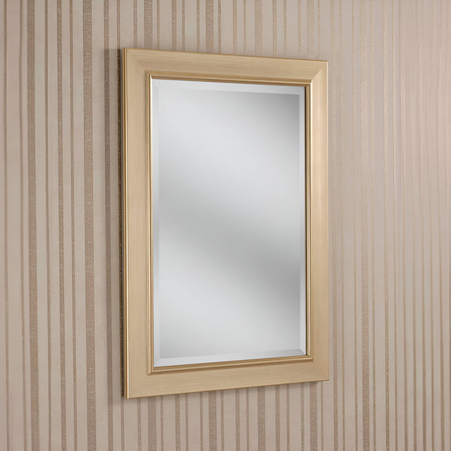 The Rimini Rectangle Wall Mirror - Gold, featuring its luxurious light wooden frame, is mounted on a wall decorated with vertical beige and light brown striped wallpaper. The mirror gracefully reflects a soft, neutral interior that exudes understated elegance.