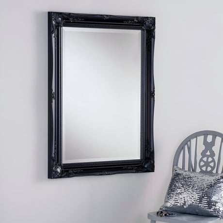 The Mirame Rectangle Wall Mirror in black, featuring an ornate frame with a modern design, is mounted on a white wall. In the lower right corner, you can partially see a gray wooden chair adorned with a metallic sequined cushion.