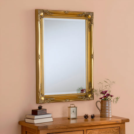 A luxurious Mirame Rectangle Wall Mirror in gold adorns the peach wall above a wooden dresser. This elegant home decor element complements the stack of books, a small clock, and a ceramic pitcher with flowers. Pinecones are quaintly scattered on the dresser.