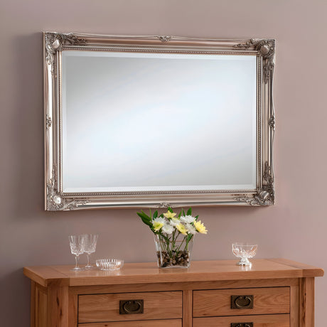 The Mirame Rectangle Wall Mirror - Silver, with its ornate frame, serves as a stunning centerpiece on the beige wall above the wooden dresser. This elegant piece of home decor perfectly complements the vase of white and yellow flowers and the two crystal glasses positioned below.