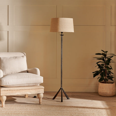Sahhil Metal Tripod Floor Lamp with antique bronze iron base and textured tapering legs, shown with a neutral linen shade beside an armchair and potted plant.
