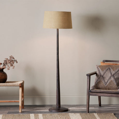 Jelvelle Mango Wood Floor Lamp with dark stained tapering column and round disc foot, shown with a tan fabric shade in a living room beside a chair and bench.