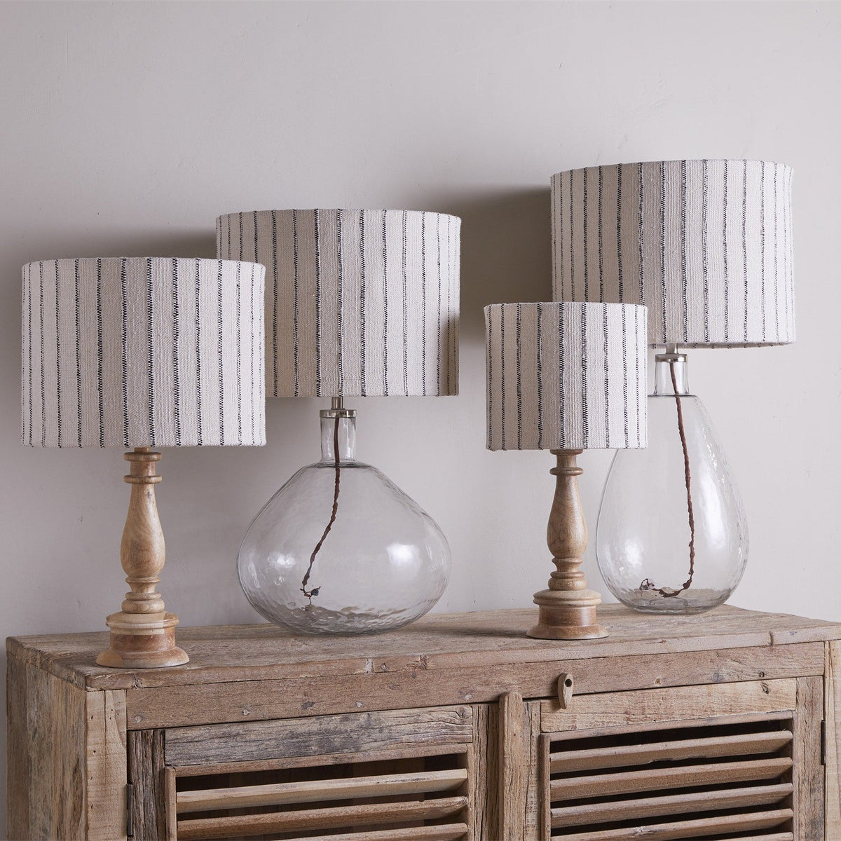 Four Sanja large cotton drum lampshades in off white with fine dark stripes, shown on wooden and glass table lamp bases atop a rustic sideboard.