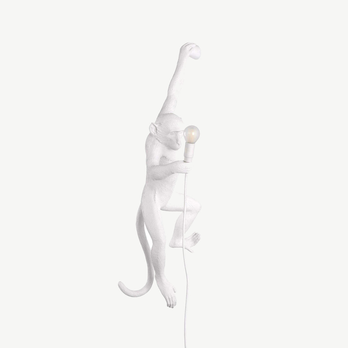 The Monkey Wall Lamp - White features a realistic monkey figure sculpted in white, climbing with one arm raised and holding a lit light bulb in the other while its tail curves downward. An electric cord accentuates the artistic design of this piece, seamlessly blending art with functionality.