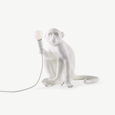 The Monkey Small Table Lamp - White features a playfully detailed white monkey figure, holding a light bulb with a trailing cord. Styled against a simple white background, this nature-inspired piece brings charm and creativity to any decor.