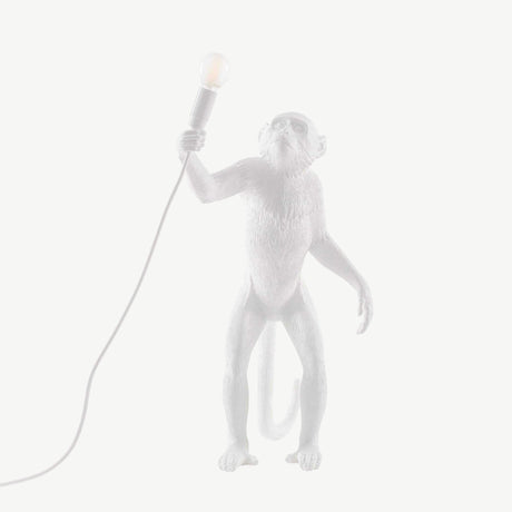 The Monkey Large Table Lamp - White features a whimsical design where an upright monkey figure holds a light bulb connected to a cord. Its minimalistic background accentuates the intricate details, transforming it into a piece of functional lighting art.