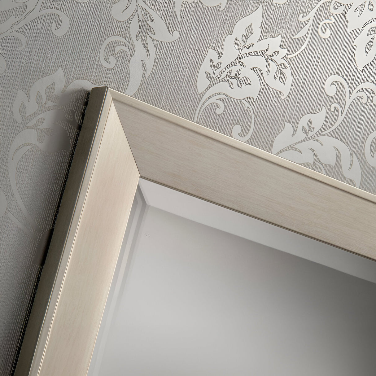 Close-up of the corner of a Sicily Rectangle Wall Mirror - Silver, featuring a wooden frame against textured wallpaper with a floral pattern. The light-colored wallpaper beautifully complements the soft silver hue of this piece, making it ideal for modern home decor.