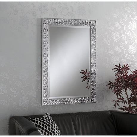 The Siena Rectangle Wall Mirror - Silver, featuring an ornate silver frame, is mounted on a patterned wall above a black leather sofa, offering a sense of modern luxury. A plant with vivid reddish leaves is positioned near the corner of the sofa, introducing a pop of color and timeless charm to the scene.