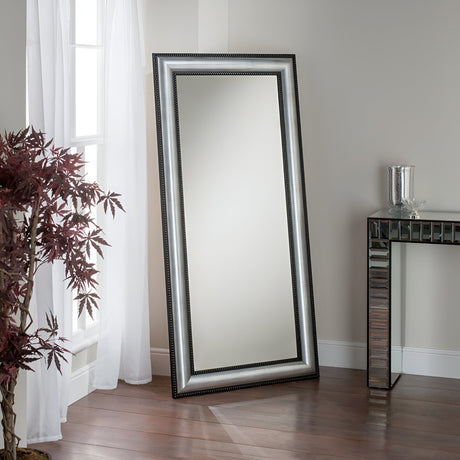 The Sorrento Leaner Mirror - Black/Silver, radiating modern sophistication, leans effortlessly against a sunlit wall. It catches the natural light streaming through white curtains, with a potted plant featuring burgundy leaves adding an earthy element on the left and a sleek glass-topped table enhancing the ambiance on the right.