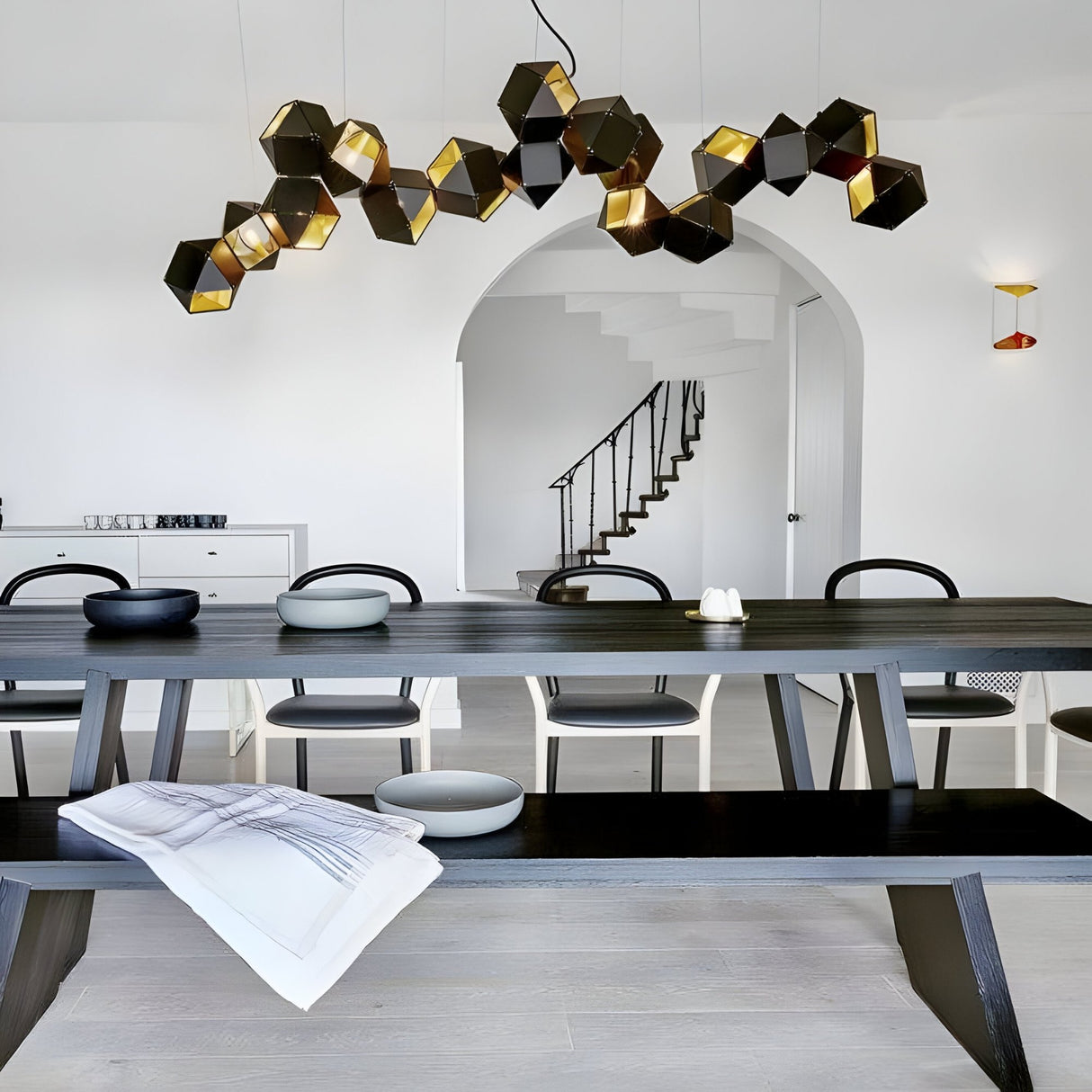 Modern dining room with a long dark wooden table, black chairs, and the striking Spar Linear Suspension Light in Black & Gold. Features white walls, an arched doorway, a staircase with black railing, and minimalist bowls on the table.
