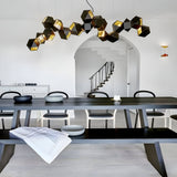 Modern dining room with a long dark wooden table, black chairs, and the striking Spar Linear Suspension Light in Black & Gold. Features white walls, an arched doorway, a staircase with black railing, and minimalist bowls on the table.