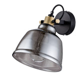 The Irving Wall Light - Black With Smoky Glass is a wall-mounted fixture featuring a black base, smoky gray glass shade, brass accents, and an exposed bulb that captures modern industrial design.