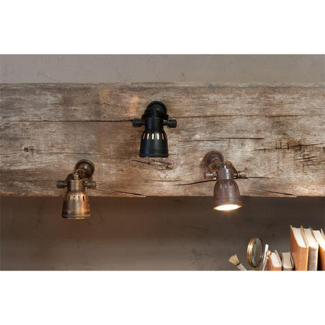 Tabosa Brass Wall Spotlight in antique brass, shown mounted on a wooden beam alongside similar dark bronze and black spotlights with knurled thumbscrews and domed slotted heads.