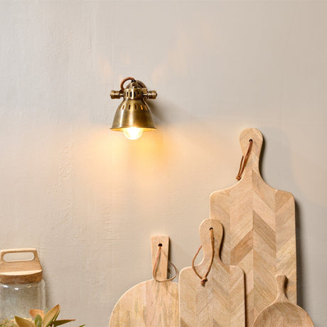 Tubu Brass Wall Spotlight in antique brass with domed head and exposed bulb, mounted on a light grey wall above wooden chopping boards.