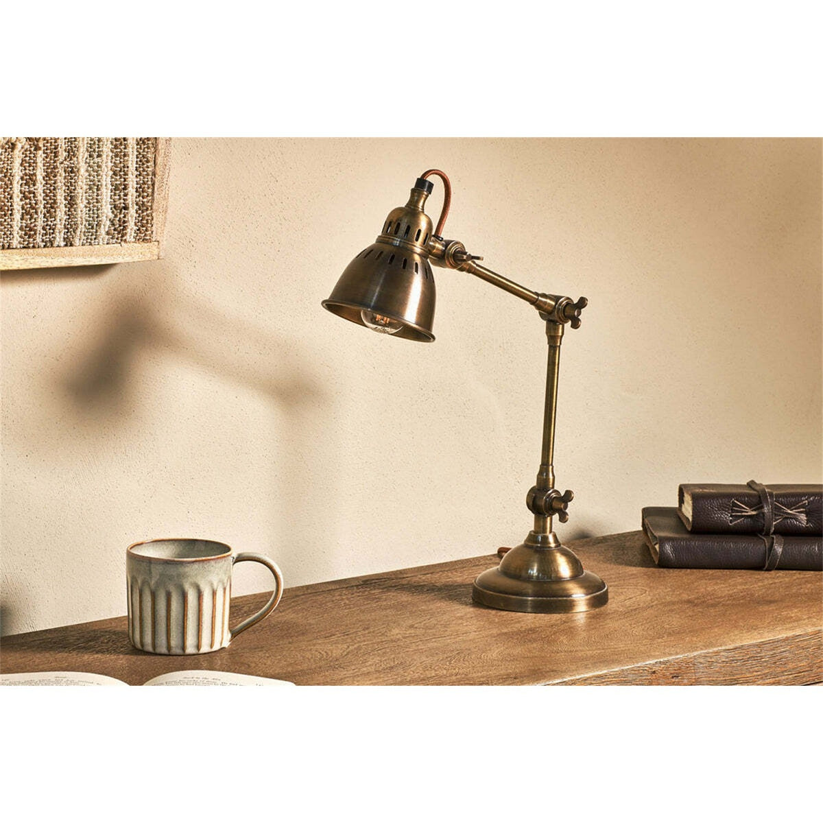 Tubu Brass Desk Lamp in antique brass with articulated arm, winged screw joints and slotted bell shade, shown on a wooden desk beside a mug and stacked books.