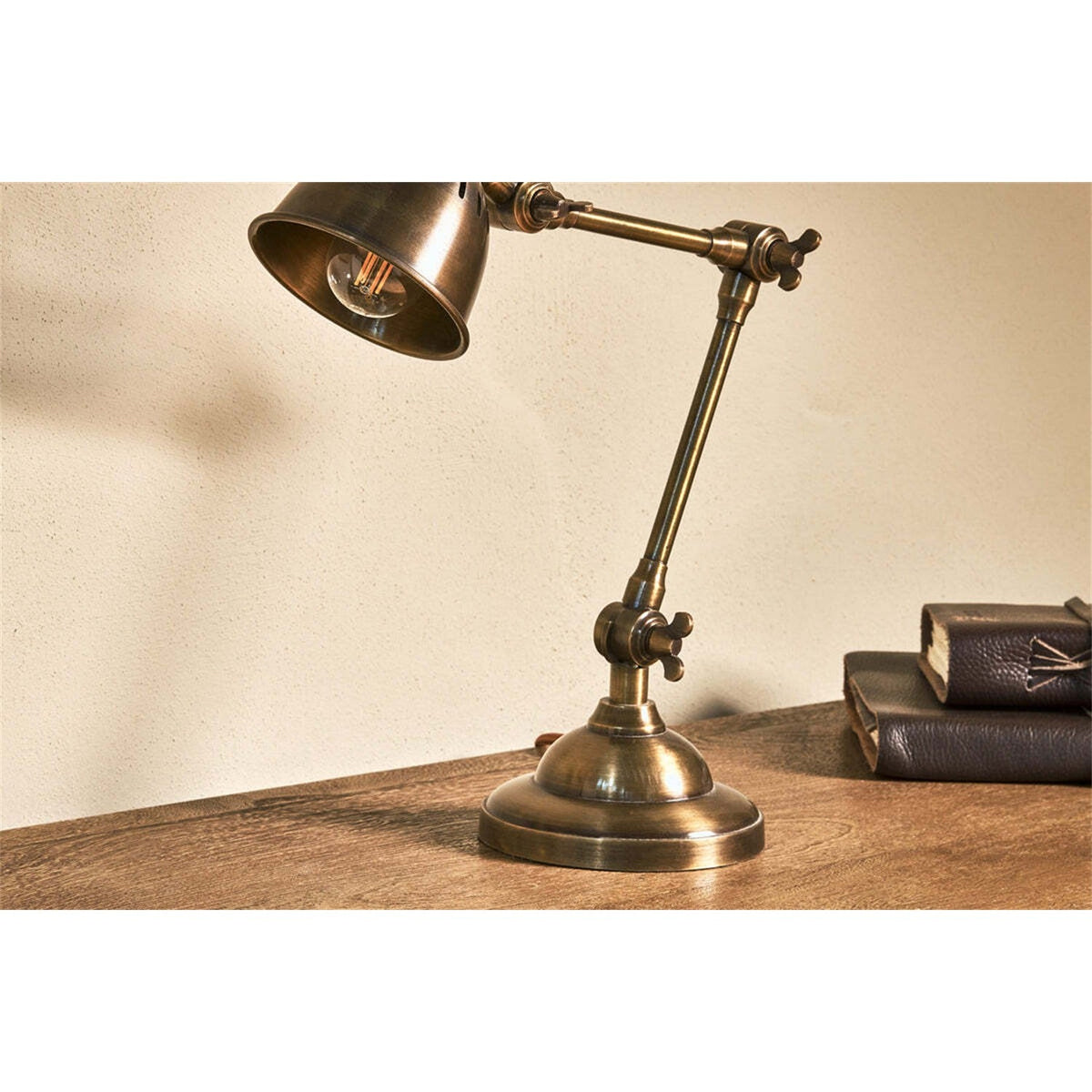 Tubu Brass Desk Lamp in antique brass with articulated arm, winged screw joints, and bell-shaped slotted shade, shown on a wooden desk beside stacked books.