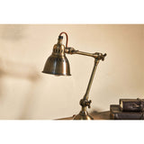 Tubu Brass Desk Lamp in antique brass, shown on a desk with an adjustable two-arm joint, winged screws, slotted bell shade, and braided flex, beside stacked leather books.