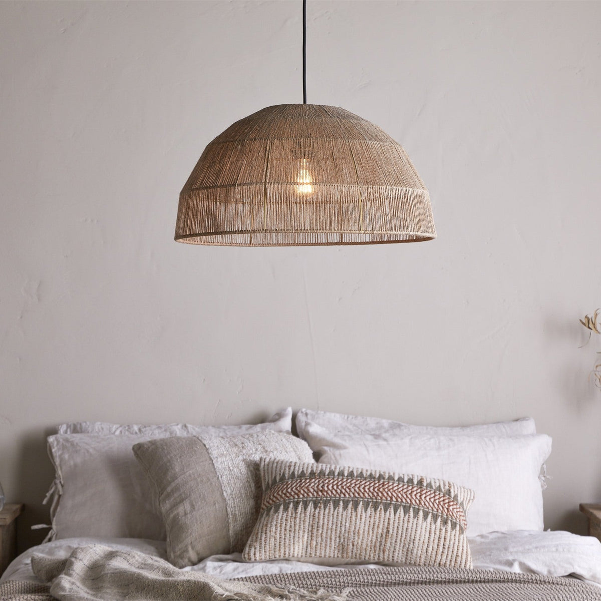 Tivi Natural Jute Lampshade, a wide dome pendant shade in woven natural jute with horizontal binding bands, hanging above a bed with linen cushions in a bedroom setting.