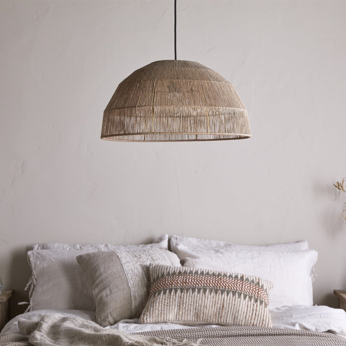 Tivi Natural Jute Lampshade, a wide dome pendant shade in woven jute cord with horizontal bands, hung above a bed with linen pillows in a neutral bedroom.