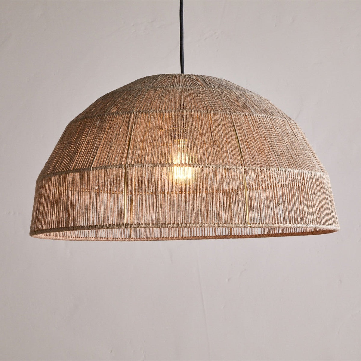 Tivi Natural Jute Lampshade, a wide dome pendant shade with vertical jute cord bound by horizontal bands, hung from a black cord against a plain light grey background, light bulb glowing inside.