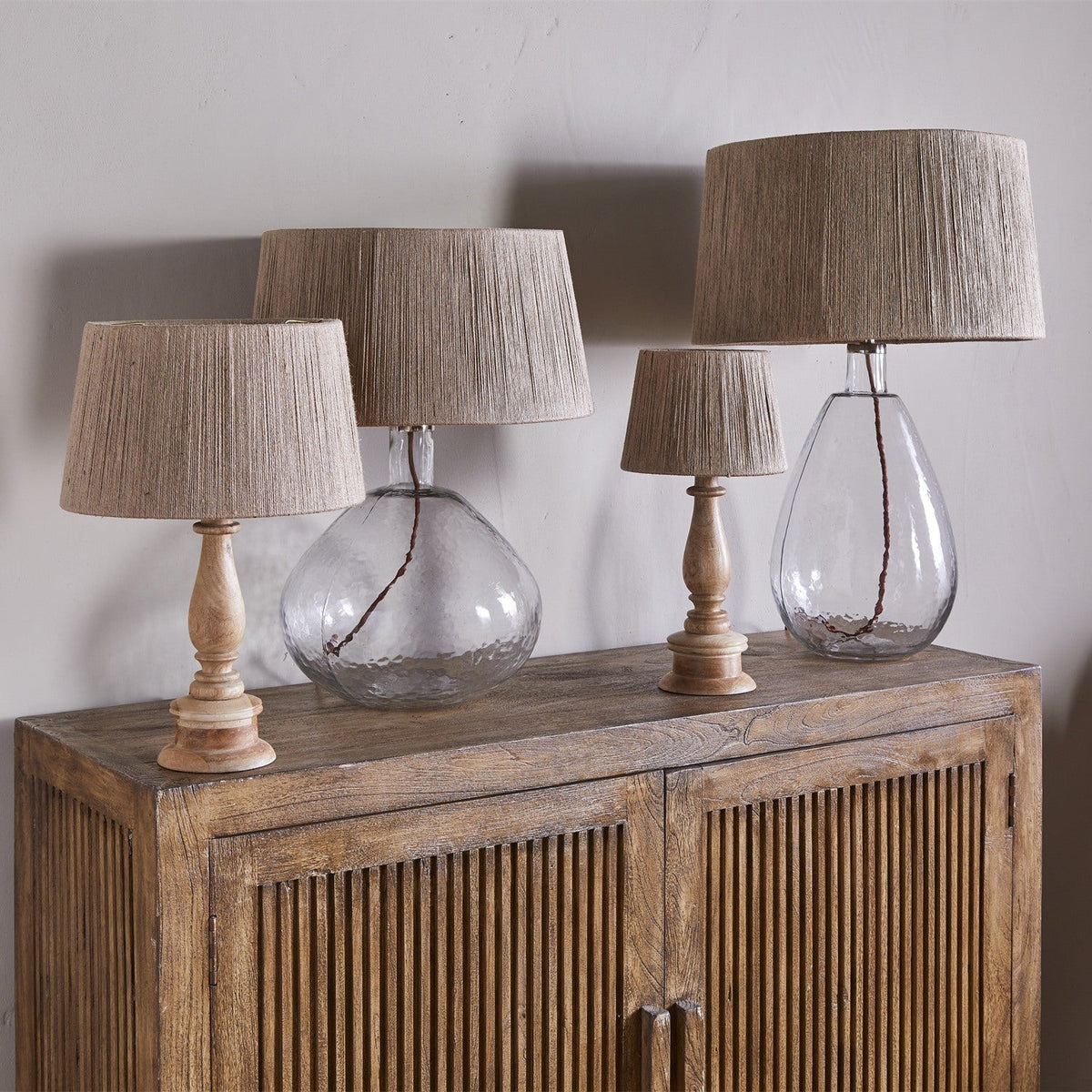 Four table lamps with tapered jute lampshades, including the Tiwa Small Woven Jute Lampshade in natural, atop turned wood and glass bases on a wooden sideboard against a plain wall.