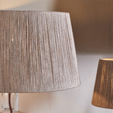 Close-up of the Tiwa Small Woven Jute Lampshade in natural straw, showing tapered drum shape with fine vertical cord lines and brass fitting ring, a second shade visible softly lit in the background.