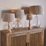 Four table lamps with Tiwa woven jute lampshades in natural tones, paired with clear glass and turned wood bases, displayed lit on a wooden sideboard.