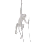 The Monkey 1 Light Pendant - White features a sculptural white monkey hanging from a rope with one hand and holding a lit lightbulb in the other, creating a striking accent against any plain backdrop.