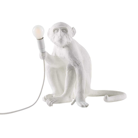 The Monkey Sitting Table Lamp - White features a sculptural white monkey holding a light bulb in its right hand, with a power cord at the base—a playful, nature-inspired lighting accent.