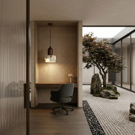 A modern minimalist workspace with a desk and chair set in an alcove features the Terroso 1 Light Drum Travertine Single Pendant Light in Walnut Brown & Light Beige. Glass walls reveal a serene Japanese-inspired zen garden with rocks, gravel, and a tree.