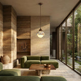 A modern living room with green velvet sofas, a wooden coffee table, and the Terroso 1 Light Dome Travertine Single Pendant Light in Walnut Brown & Light Beige. Large floor-to-ceiling windows reveal lush greenery beyond beige stone walls.