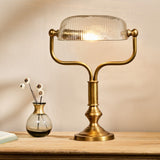 Ulani Vintage Desk Lamp in antique brass with a ribbed clear glass dome shade on a twin-stem yoke and stepped round base, shown lit on a wooden surface beside a small vase.