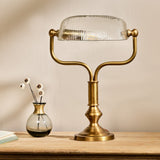 Ulani Vintage Desk Lamp in antique brass with a ribbed clear glass dome shade, twin-stem yoke and stepped round base, shown on a wooden surface beside a small glass vase.