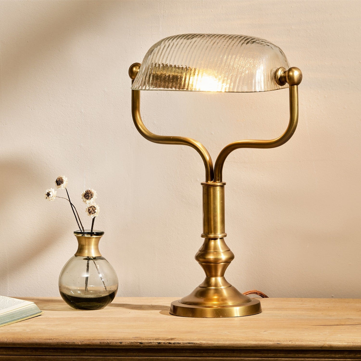 Ulani Vintage Desk Lamp in antique brass with ribbed clear glass banker-style dome shade, shown lit on a wooden table beside a small vase.