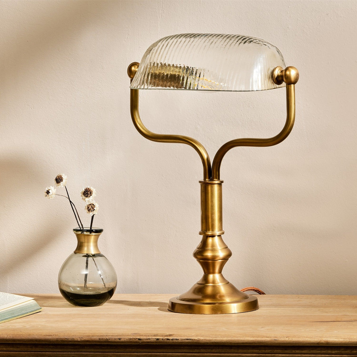 Ulani Vintage Desk Lamp in antique brass with a ribbed clear glass banker-style shade, shown on a wooden surface beside a small brass vase.