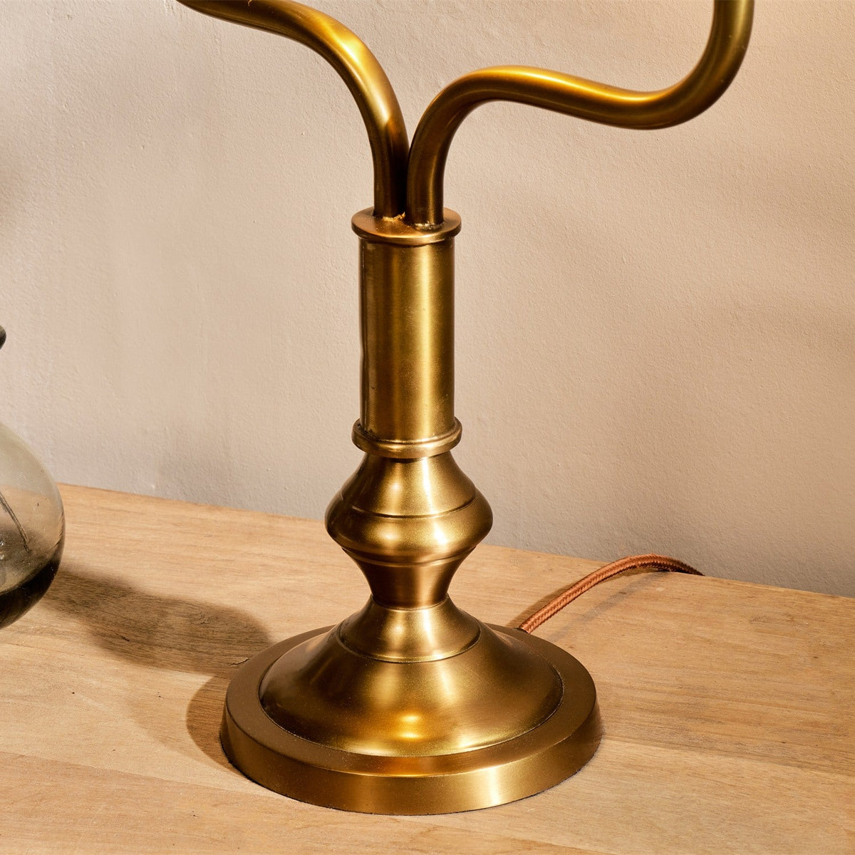 Close-up of the Ulani Vintage Desk Lamp's antique brass column and turned base with twin stem yoke, shown on a wooden surface against a plain wall.