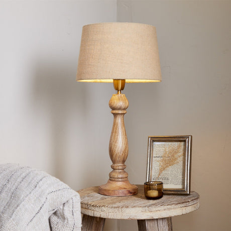Usha Large Classic Wooden Table Lamp in mango wood with baluster-turned column, brass collar, and tapered beige fabric shade, shown on a rustic side table.