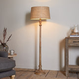 Usha Classic Wooden Floor Lamp with turned mango wood candlestick base in natural finish, shown with a taupe drum shade in a living room setting.