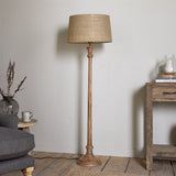 Usha Classic Wooden Floor Lamp with turned mango wood candlestick base and natural beige drum shade, shown beside a console table in a living room setting.