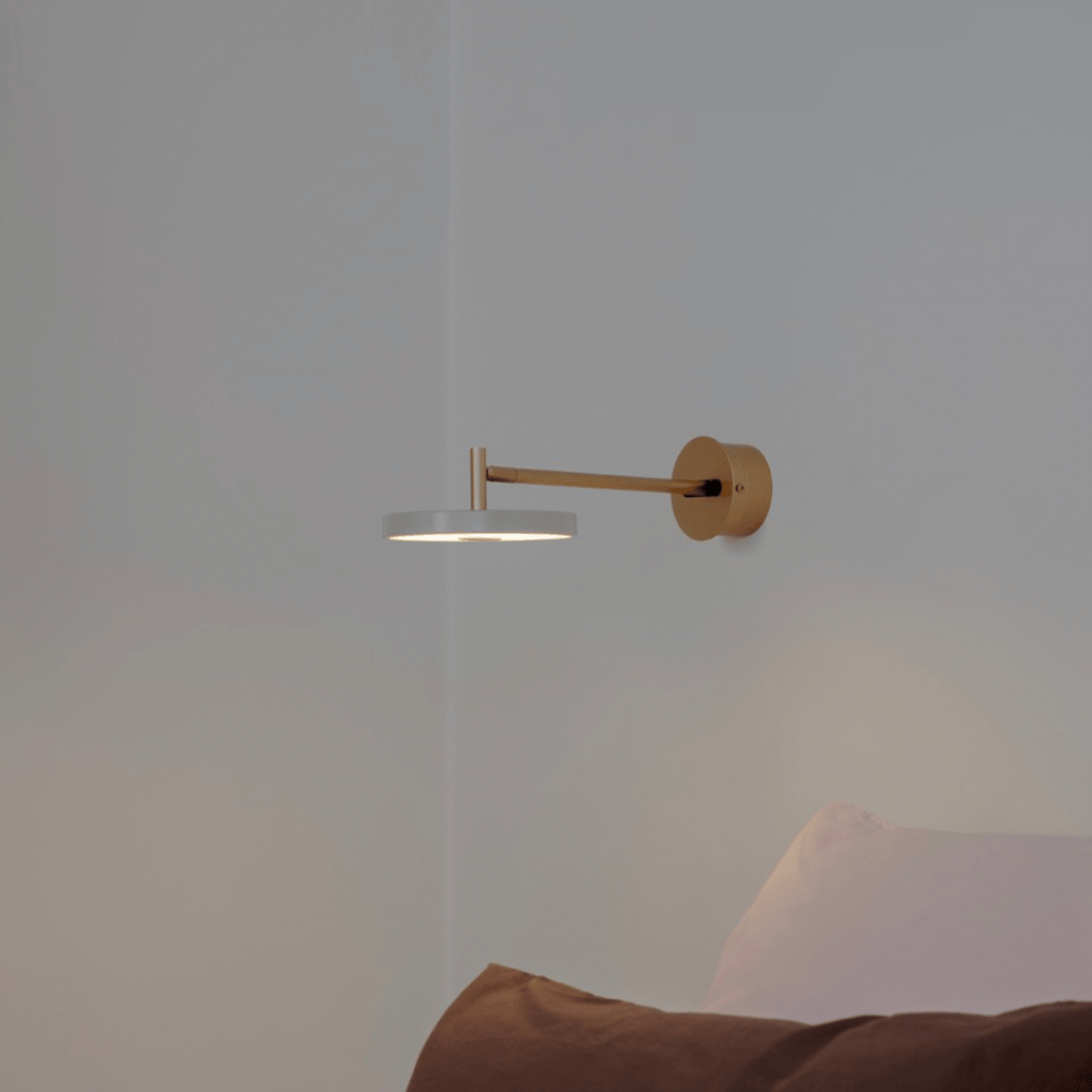 Asteria Long LED Wall Light in Brass with a Nuance Mist disc shade, long straight arm from a round wall plate, shown lit above a bed in a bedroom setting.