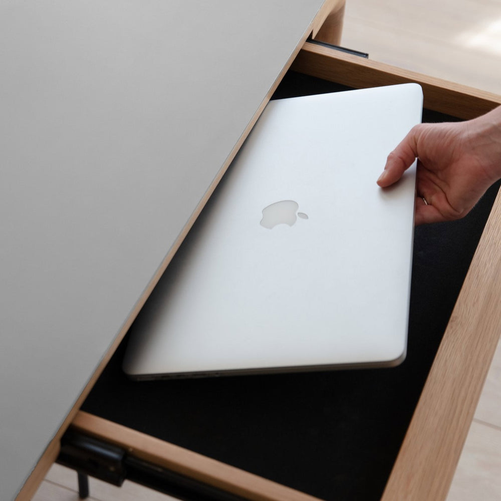 Open drawer with soft black lining beneath a Cloud Grey tabletop, shown as part of the Heart'n'Soul 120 Extendable Oak Dining Table, with a hand placing a laptop inside.