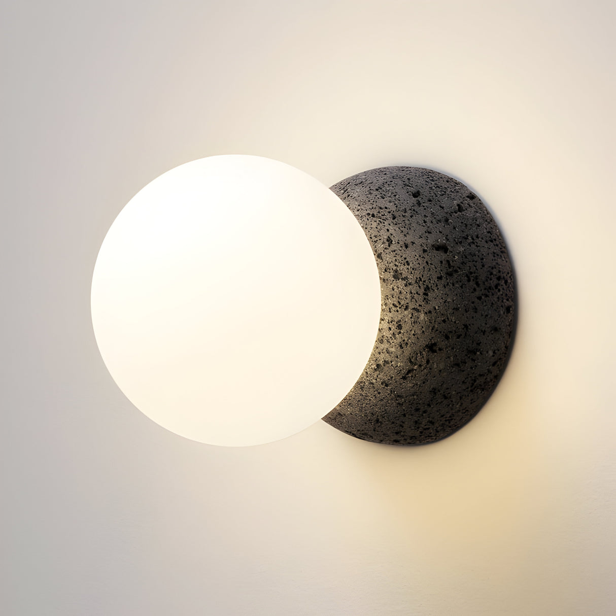 The Origo Concrete Wall Light - Black & White features a wall-mounted design with a spherical white bulb and a base resembling volcanic rock. Its opal glass diffuser enhances illumination, casting a soft glow on the wall and highlighting the fixture's simple, modern aesthetic.
