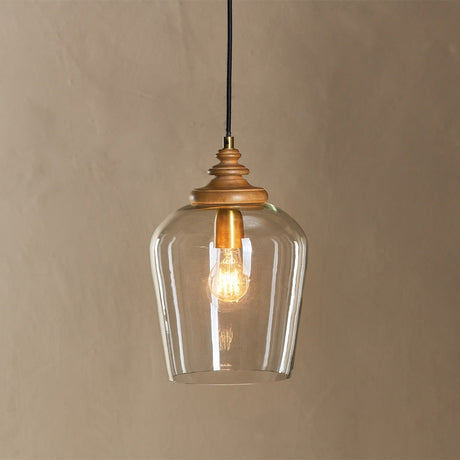 Varuna pendant light with a clear glass bell shade on a turned mango wood collar, brass ring and fittings, black fabric flex, exposed bulb, neutral background.