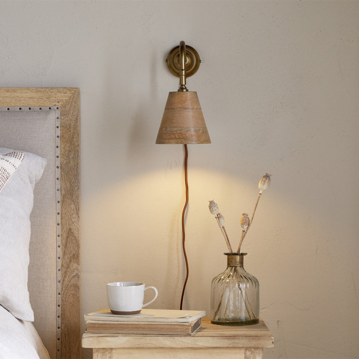 Vattu plug-in wall light with brass wall plate, swan neck arm, and mango wood cone shade, shown lit beside a bed with a nightstand, mug, and vase.