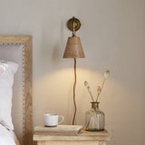 Vattu plug-in wall light with brass wall plate, swan neck arm, and mango wood cone shade, shown lit beside a bed with a nightstand, mug, and vase.