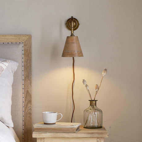 Vattu plug-in wall light with brass wall plate, swan neck arm, and mango wood cone shade, shown lit beside a bed with a nightstand, mug, and vase.