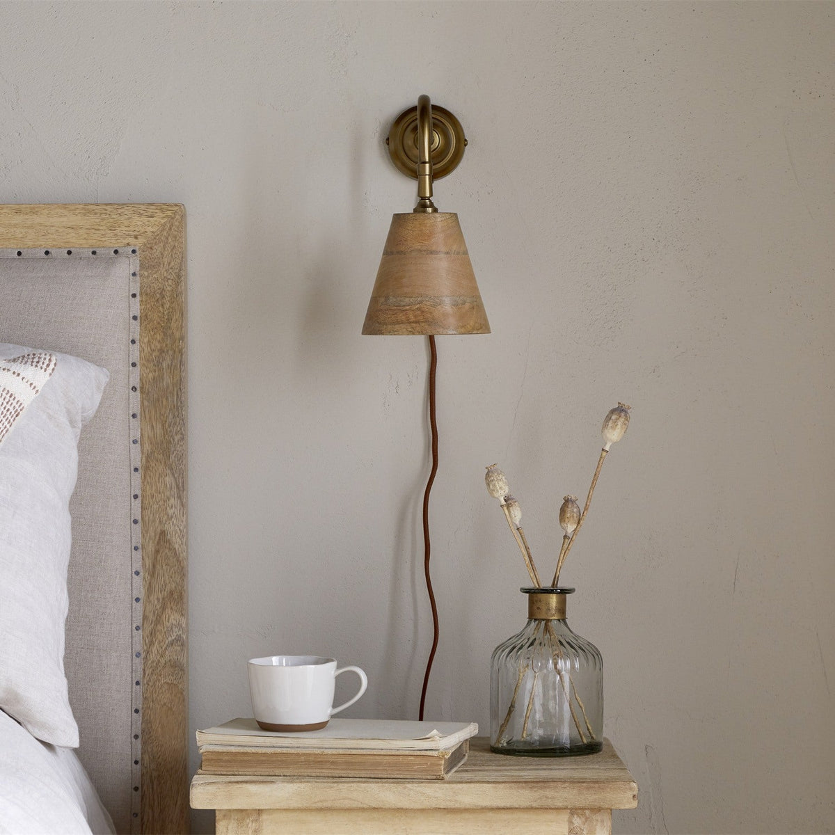 Vattu Mango Wood Plug In Wall Light with a brass wall plate and swan neck arm, cone-shaped mango wood shade and rust-coloured cord, shown above a bedside table in a bedroom setting.