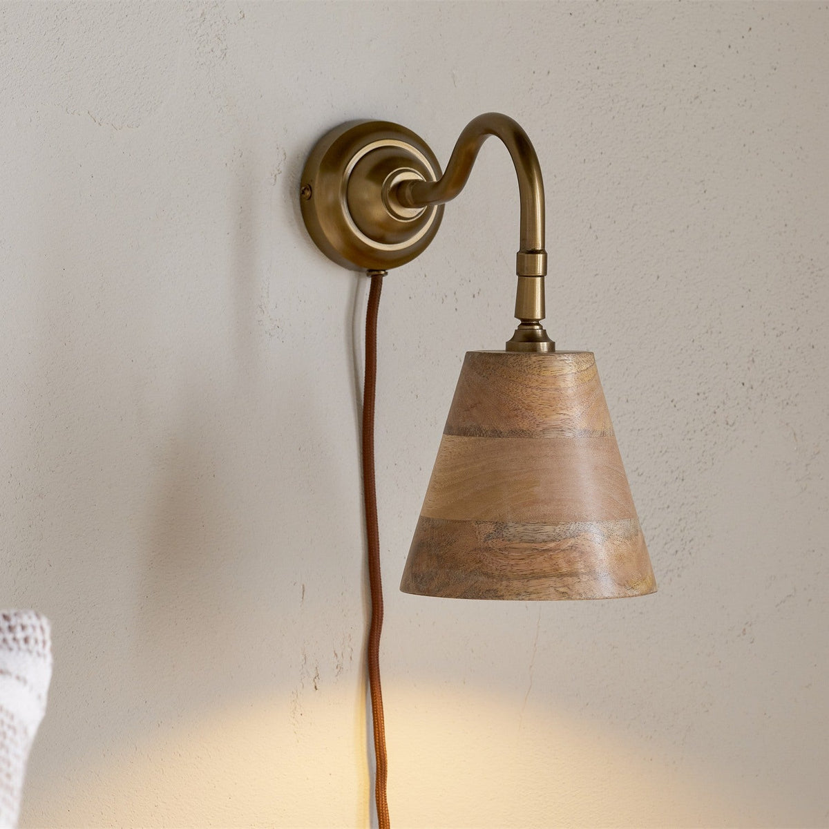 Vattu plug-in wall light with antique brass swan neck arm and round wall plate, mango wood cone shade in natural honey tones, rust-coloured cord, shown mounted on a plain wall.