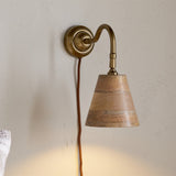 Vattu plug-in wall light with antique brass swan neck arm and round wall plate, mango wood cone shade in natural honey tones, rust-coloured cord, shown mounted on a plain wall.