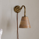 Vattu Mango Wood Plug In Wall Light in Natural, featuring an antique brass wall plate and swan neck arm with a mango wood cone shade and rust-coloured cord, shown against a textured light wall.